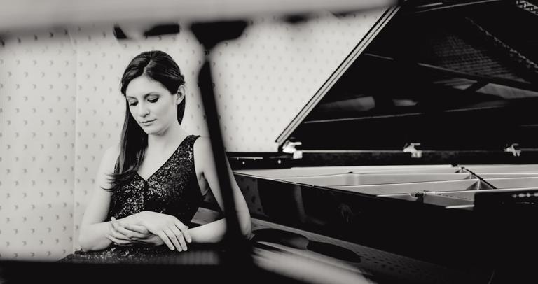 Eine junge Frau mit langen, dunklen Haaren und geschlossenen Augen sitzt rückwärts zum Instrument an einem offenen Flügel, wobei sie einen Ellenbogen auf den geschlossenen Tastaturkasten abstützt.