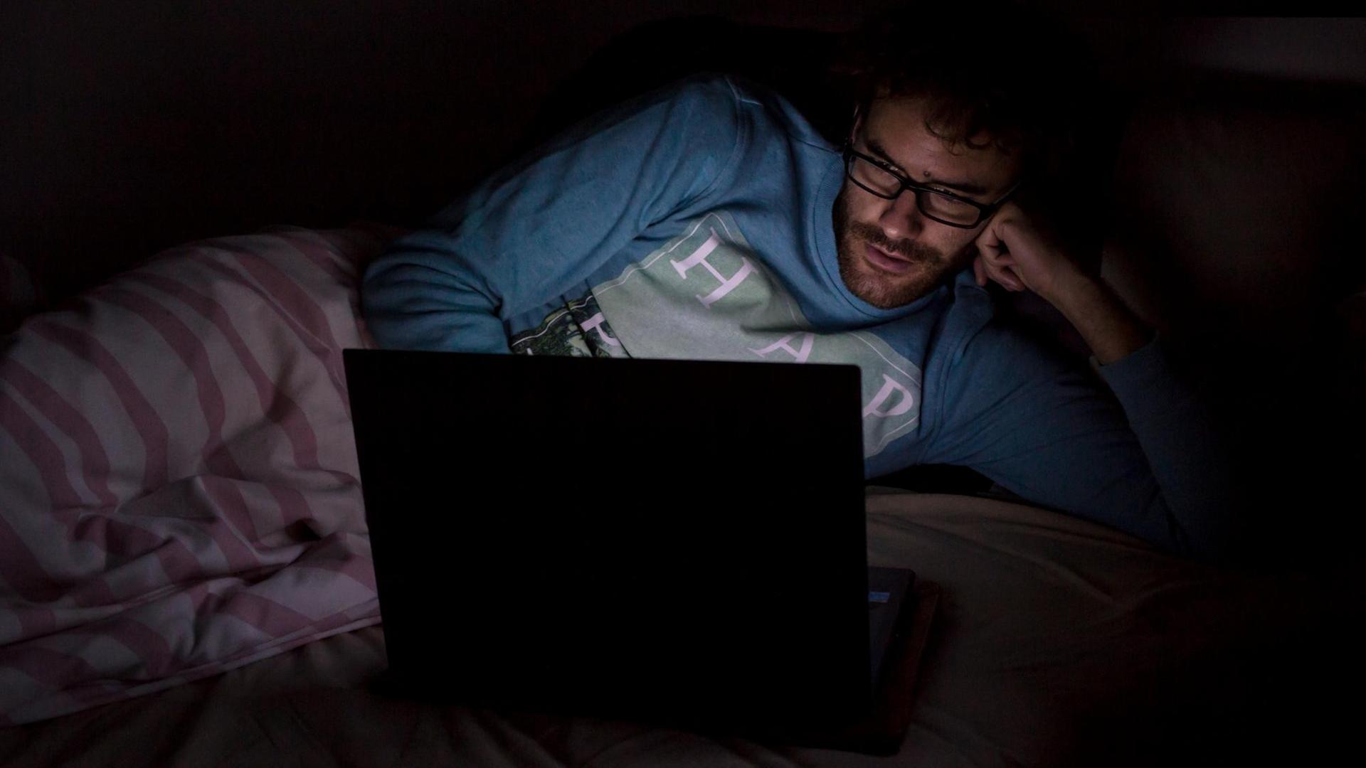 Ein Mann liegt im Dunkeln auf seinem Bett und schaut auf den Monitor eines aufgeklappten Laptops. Ein Mann liegt im Dunkeln auf seinem Bett und schaut auf den Monitor eines aufgeklappten Laptops.