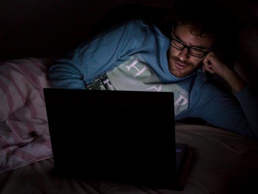 Ein Mann liegt im Dunkeln auf seinem Bett und schaut auf den Monitor eines aufgeklappten Laptops.