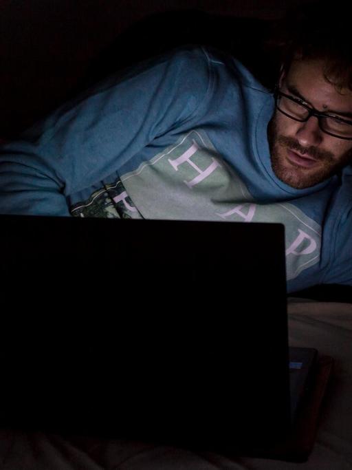 Ein Mann liegt im Dunkeln auf seinem Bett und schaut auf den Monitor eines aufgeklappten Laptops.