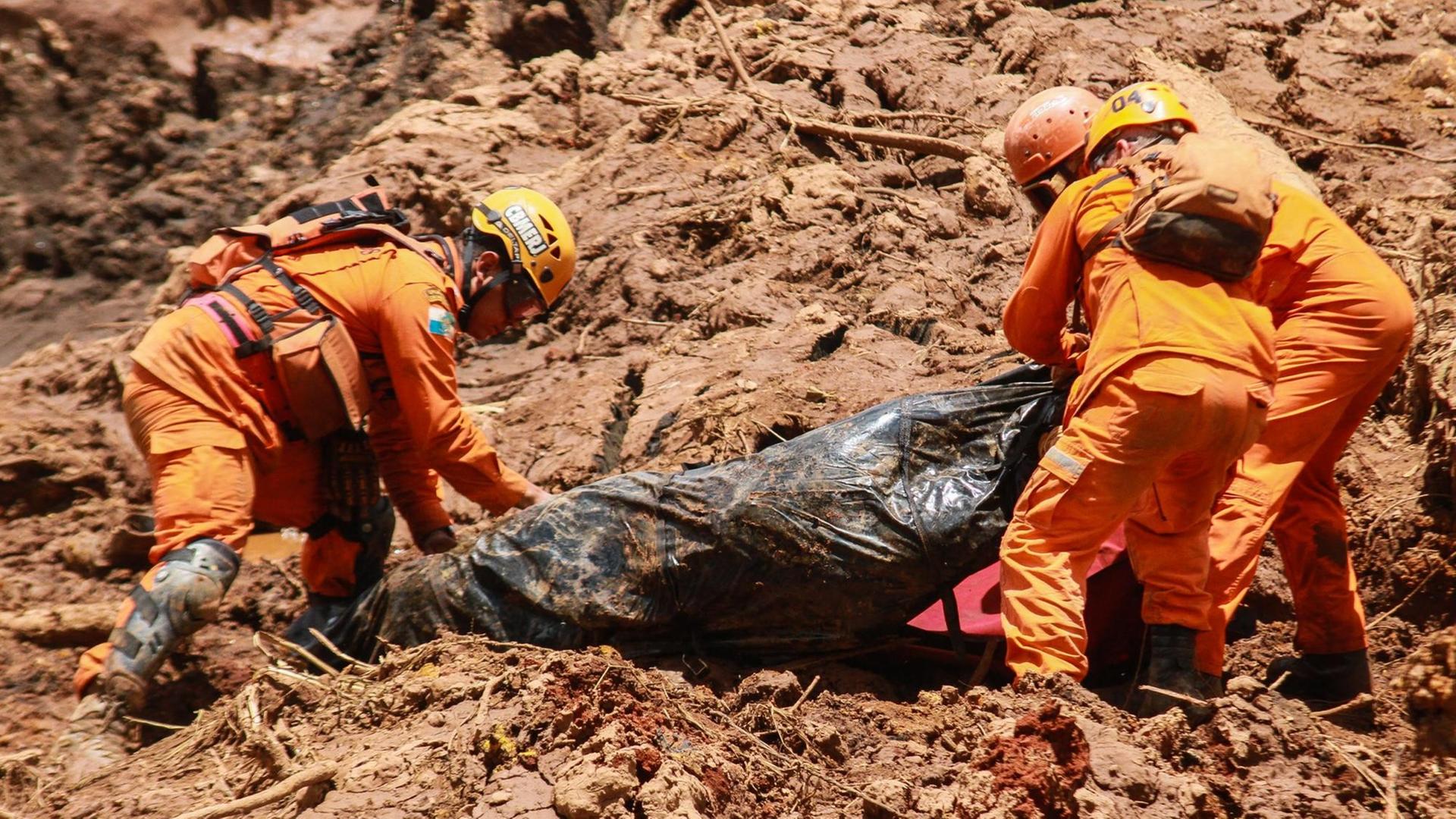 Die drei Männer in Helmen und orangefarbener Arbeitskleidung ziehen in der braunen Schlammwüste an einem Leichensack. Die drei Männer in Helmen und orangefarbener Arbeitskleidung ziehen in der braunen Schlammwüste an einem Leichensack.