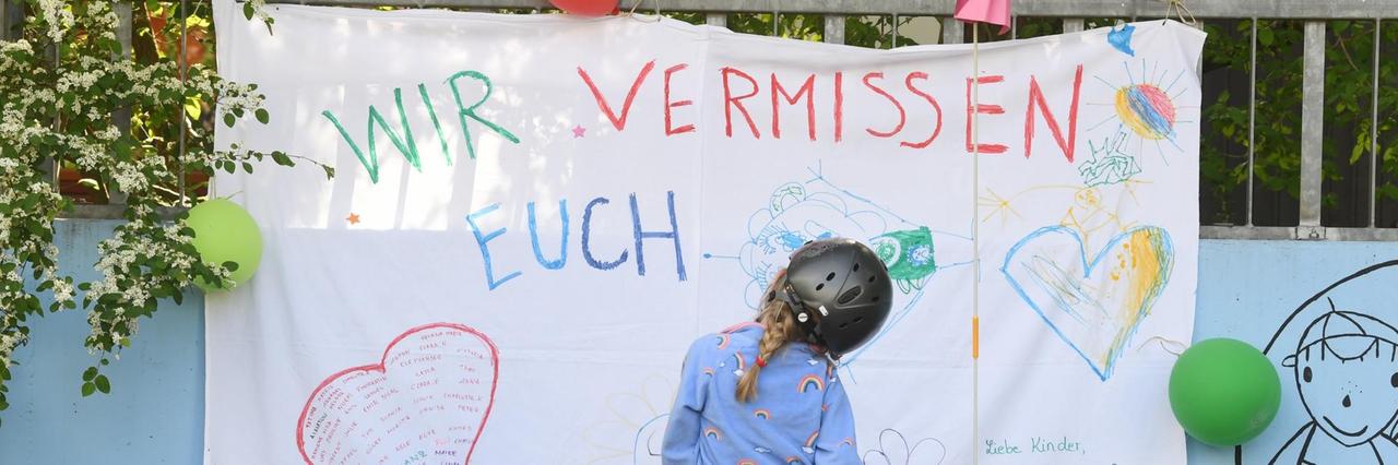 Ein Mädchen auf dem Fahrrad steht vor der Fassade einer Kita mit der Aufschrift "Wir vermissen euch". Ein Mädchen auf dem Fahrrad steht vor der Fassade einer Kita mit der Aufschrift "Wir vermissen euch".