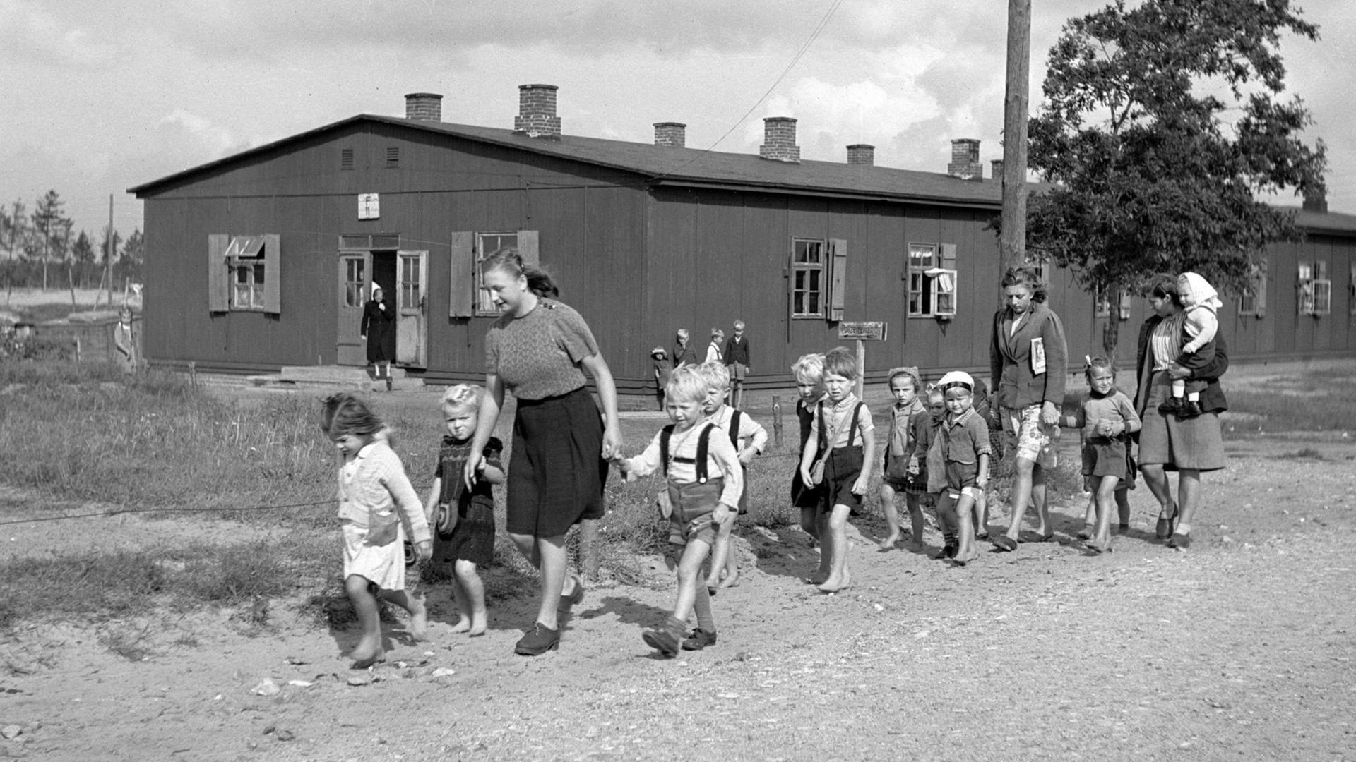 Eine Gruppe von Kindern, begleitet von zwei Frauen, läuft einen Weg entlang. Im Hintergrund sind Holzhäuser zu sehen. Eine Gruppe von Kindern, begleitet von zwei Frauen, läuft einen Weg entlang. Im Hintergrund sind Holzhäuser zu sehen.