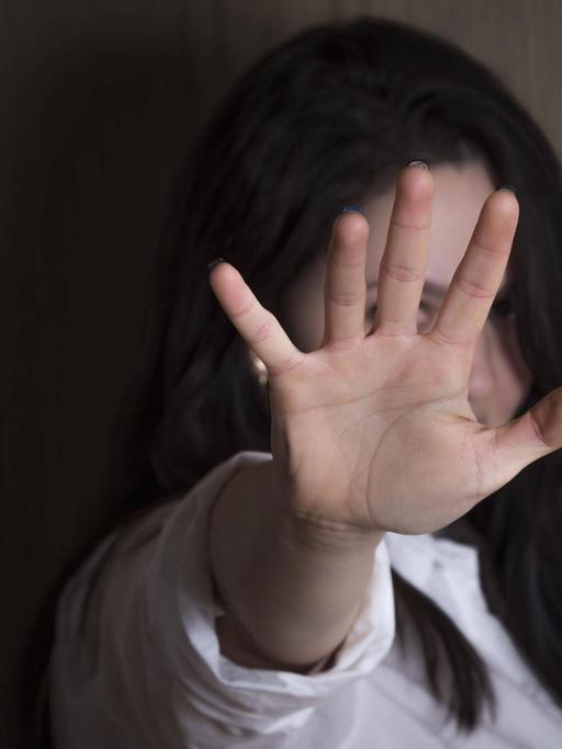 Eine Frau vor einer Holzwand hat ihre Hand zu einer Stopp-Geste ausgestreckt, und verdeckt so ihr Gesicht.