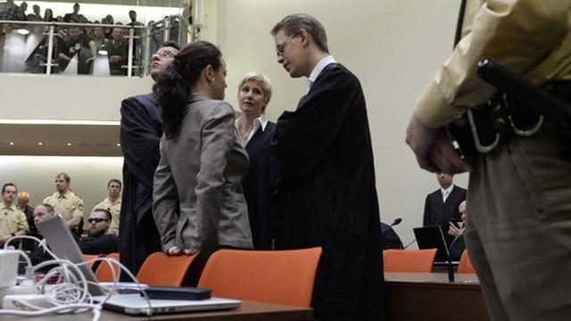 Beate Zschäpe steht im Münchner Gerichtssaal an einen Stuhl gelehnt, umringt von ihren Anwälten.