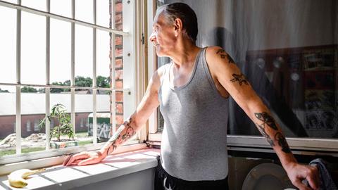 Helmut, graue zurückgekämmte Haare, graues Tanktop, Tattoos auf den Armen, steht am geöffneten Fenster, stützt seine rechte Hand auf die Fensterbank und schaut durch die Gefängnisgitter hinaus. Die Sonne scheint.