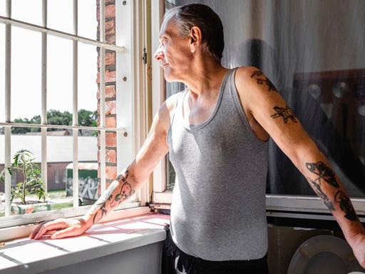 Helmut, graue zurückgekämmte Haare, graues Tanktop, Tattoos auf den Armen, steht am geöffneten Fenster, stützt seine rechte Hand auf die Fensterbank und schaut durch die Gefängnisgitter hinaus. Die Sonne scheint.