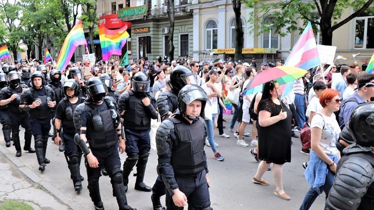 Menschen in Sommerkleidung schwenken Regenbogenflaggen, auch eine Transgender-Pride-Flagge ist im Bild, im Vordergrund zwei Reihen Polizei in voller, schwarzer Montur Menschen in Sommerkleidung schwenken Regenbogenflaggen, auch eine Transgender-Pride-Flagge ist im Bild, im Vordergrund zwei Reihen Polizei in voller, schwarzer Montur