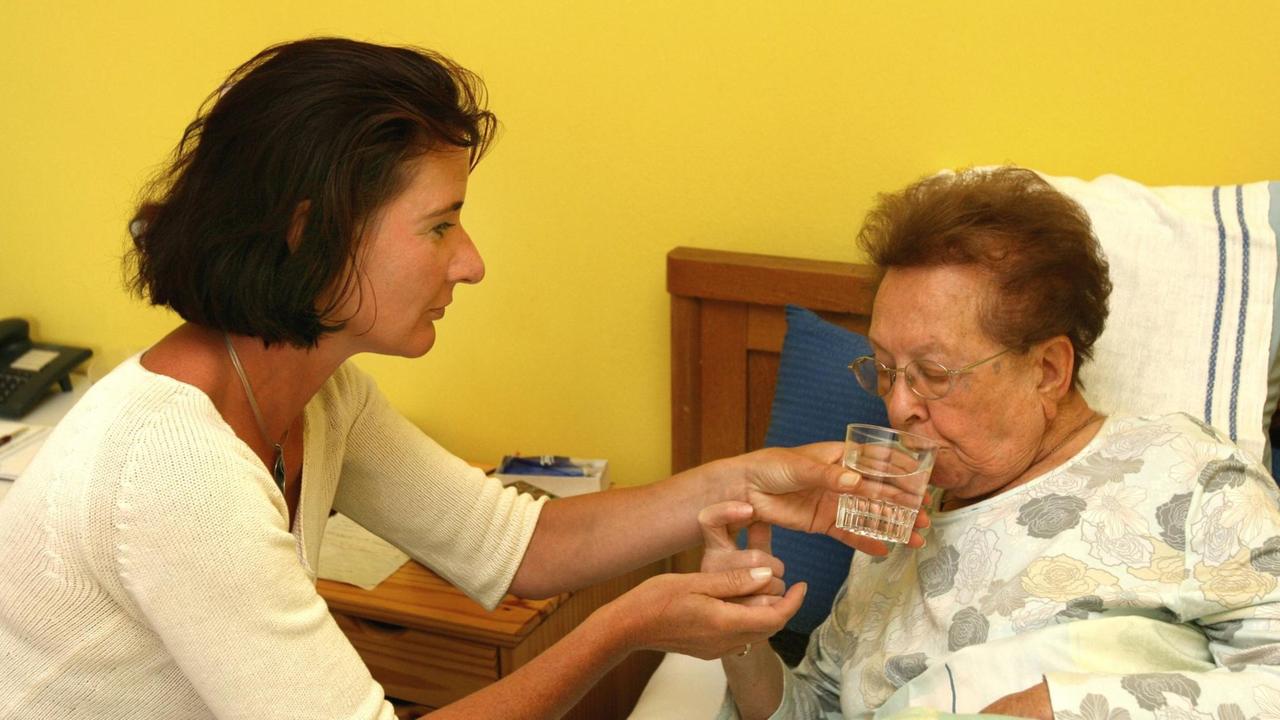 Eine Pflegerin reicht einer bettlägerigen Seniorin ein Glas Wasser. Eine Pflegerin reicht einer bettlägerigen Seniorin ein Glas Wasser.