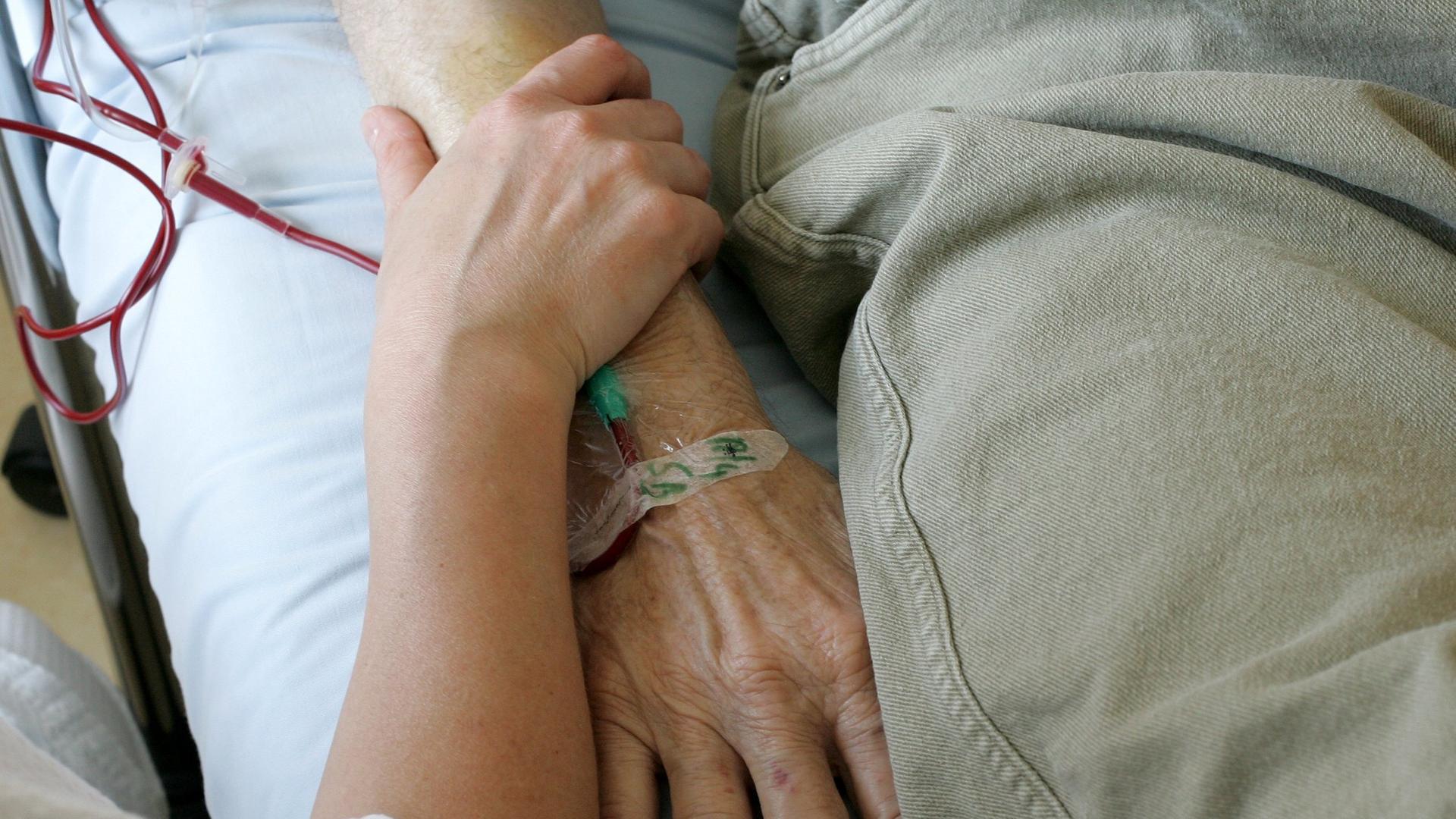 Ein jüngerer Mensch umfasst das Armgelenk einer älteren Person, die im Krankenbett liegt. Ein jüngerer Mensch umfasst das Armgelenk einer älteren Person, die im Krankenbett liegt.