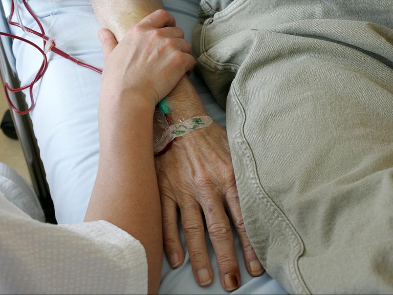 Ein jüngerer Mensch umfasst das Armgelenk einer älteren Person, die im Krankenbett liegt.