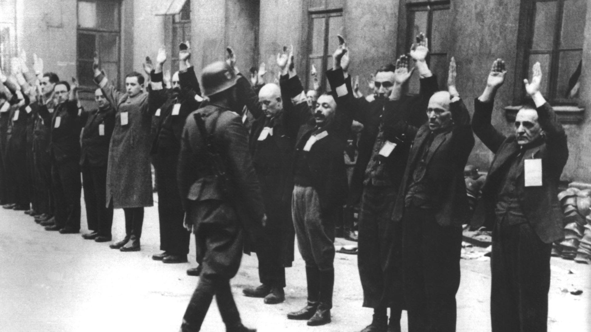 Bewacht von einem deutschen Soldaten stehen Bewohner des Warschauer Ghettos mit erhobenen Armen in einem Innenhof. Bewacht von einem deutschen Soldaten stehen Bewohner des Warschauer Ghettos mit erhobenen Armen in einem Innenhof.