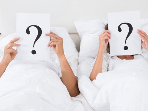 Zwei Menschen liegen in einem Bett und halten jeweils ein Blatt mit einem Fragezeichen darauf vor ihre Gesichter.