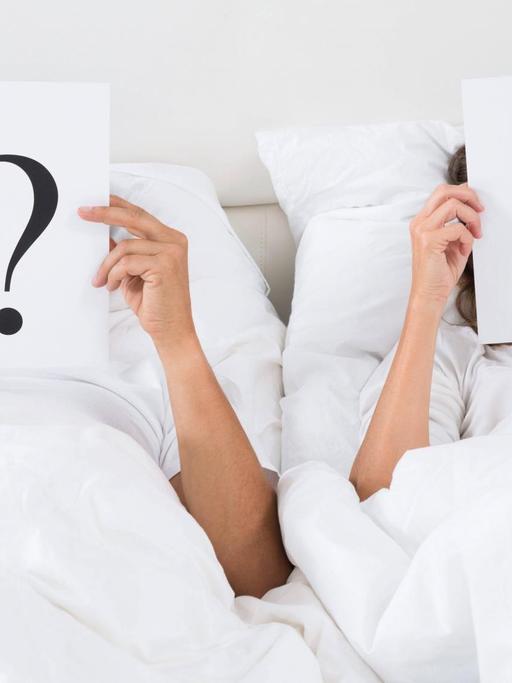 Zwei Menschen liegen in einem Bett und halten jeweils ein Blatt mit einem Fragezeichen darauf vor ihre Gesichter.