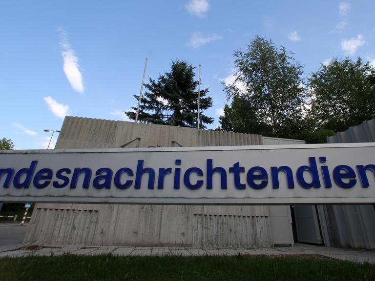 Graues Schild mit blauer Aufschrift "Bundesnachrichtendienst" vor einer grauen Mauer und blauem Himmel