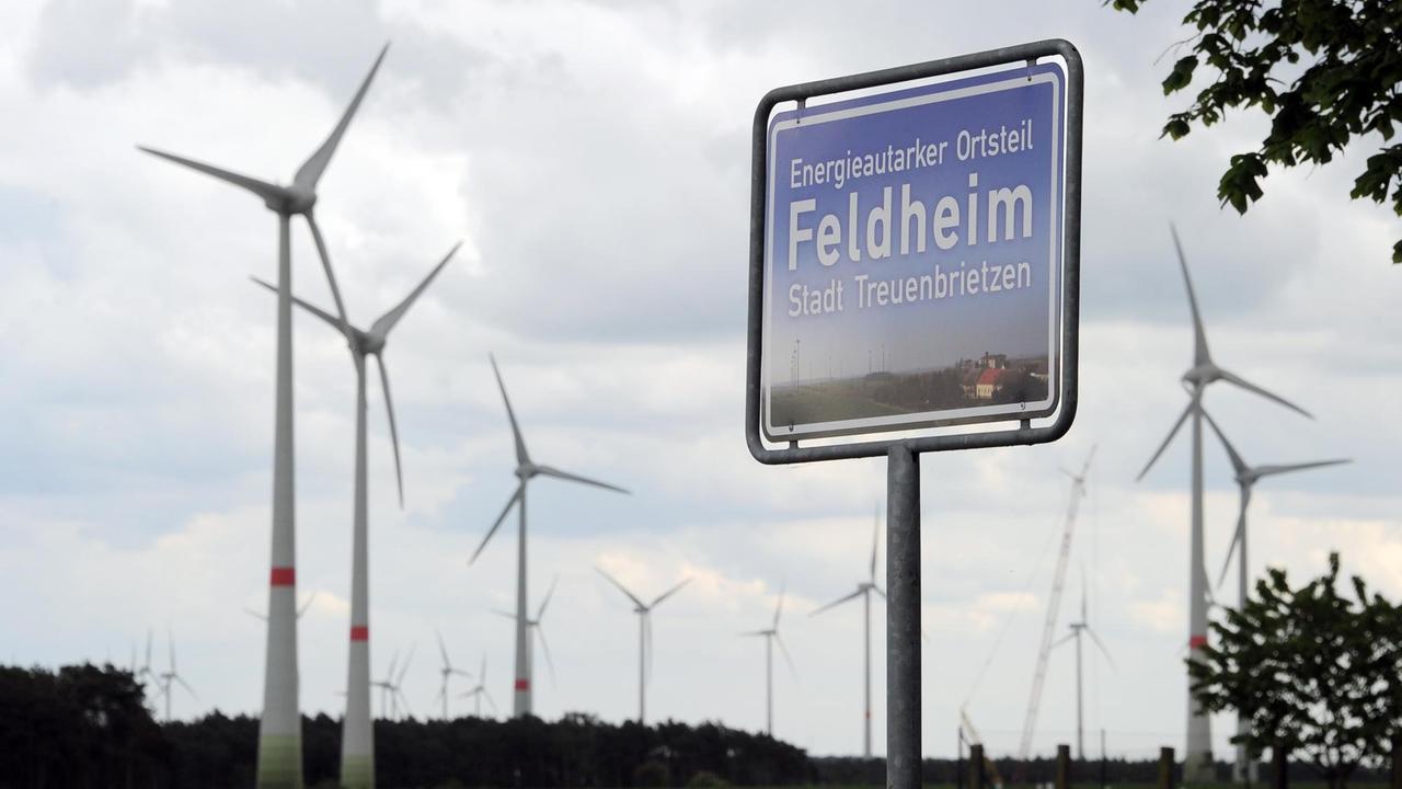 Ein blaues Schild, hinter dem sich Windräder drehen, weist Feldheim (Brandenburg) als energieautarken Ort aus. 