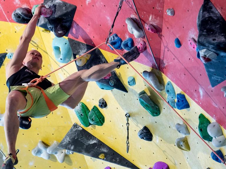 Christoph Hanke klettert im März 2018 mit einem Seil gesichert an einer Kletterwand. Sein rechtes Bein hängt in der Luft, mit den übrigen Extremitäten hält er sich fest und stützt sich ab.