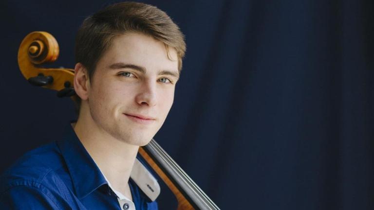 Ein junger Mann mit kurzen, hellbraunen Haaren schaut in einer Porträtaufnahme in die Kamera, an seiner linken Schulter lehnt der Hals eines Cellos. Ein junger Mann mit kurzen, hellbraunen Haaren schaut in einer Porträtaufnahme in die Kamera, an seiner linken Schulter lehnt der Hals eines Cellos.