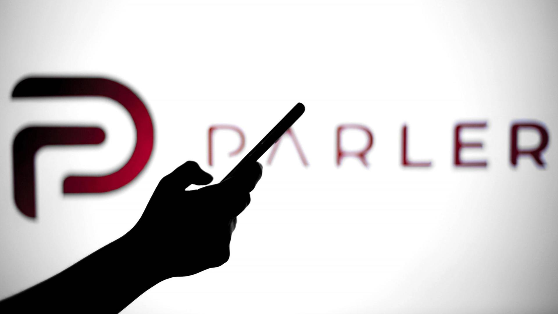 Eine Hand hält ein Smartphone, hinter dem Umriss ist vor leuchtend weißem Hintergrund das Logo von "Parler" zu sehen. Eine Hand hält ein Smartphone, hinter dem Umriss ist vor leuchtend weißem Hintergrund das Logo von "Parler" zu sehen.