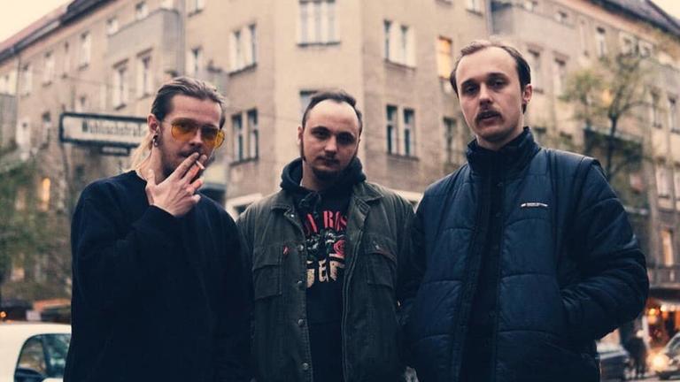 Die drei Musiker von Molchat Doma stehen nebeneinander, hinter ihnen befindet sich ein Berliner Altbau.