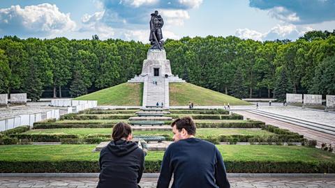 Eine junge Frau und ein junger Mann sitzen nebeneinander, im Hintergrund ist das Sowjetische Ehrenmal im Treptower Park in Berlin zu sehen.
