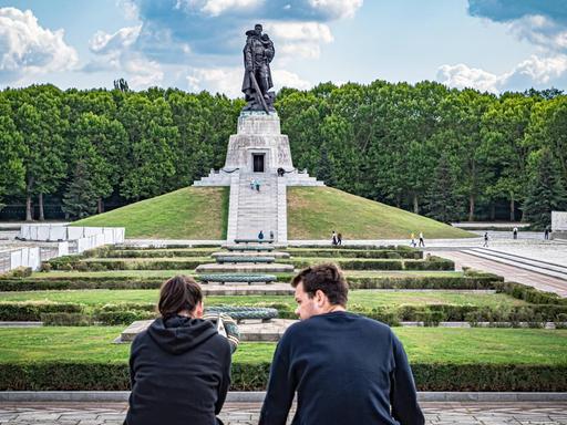 Eine junge Frau und ein junger Mann sitzen nebeneinander, im Hintergrund ist das Sowjetische Ehrenmal im Treptower Park in Berlin zu sehen.