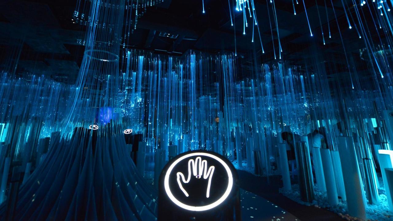 Eine interaktive Kunstinstallation aus blau leuchteden Stäben, im Bildvordergrund ist ein Leuchtzeichen mit den Umrissen einer Hand zu sehen. Eine interaktive Kunstinstallation aus blau leuchteden Stäben, im Bildvordergrund ist ein Leuchtzeichen mit den Umrissen einer Hand zu sehen.
