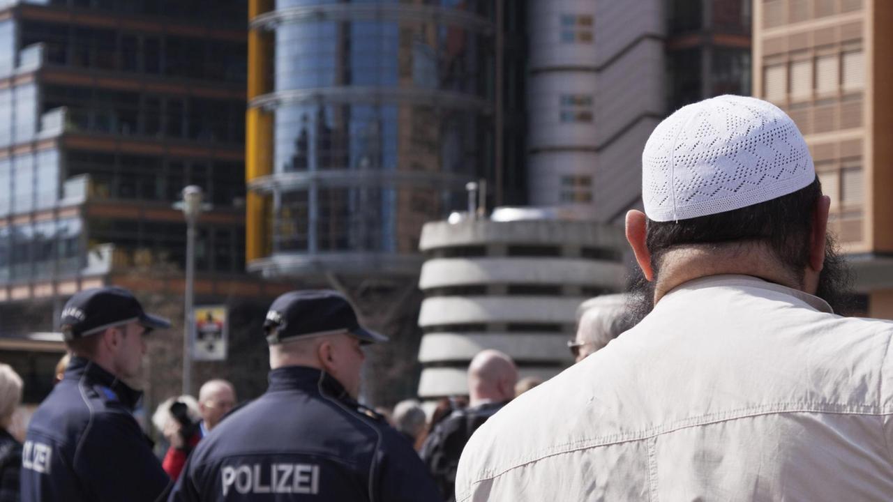 Rückenansicht eines Mannes mit Gebetsmütze, der in Richtung zweier uniformierter Polizisten blickt.