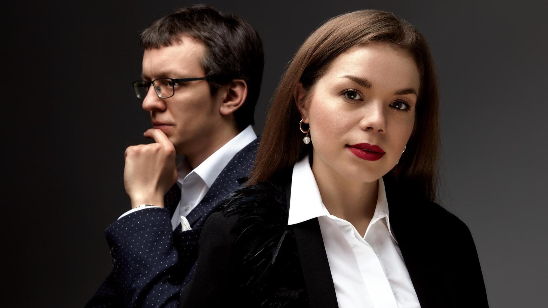 Eine Frau in weißer Bluse und schwarzme Blazer blickt in die Kamera, sie trägt auffallenden, roten Lippenstift und hält den Kopf leicht geneigt. Links hinter hier steht ein Mann mit Brille und schaut nach links aus dem Bild. Eine Frau in weißer Bluse und schwarzme Blazer blickt in die Kamera, sie trägt auffallenden, roten Lippenstift und hält den Kopf leicht geneigt. Links hinter hier steht ein Mann mit Brille und schaut nach links aus dem Bild.