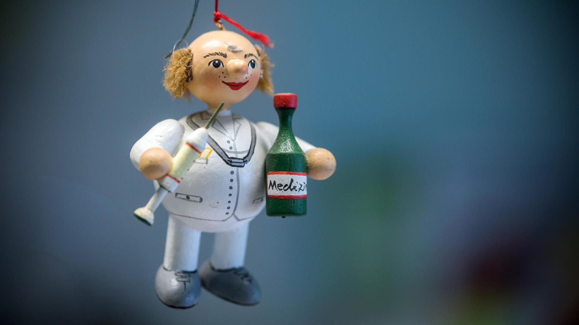 Eine Holzfigur eines Doktors mit Spritze und Medizinflächen hängt an einem roten Faden in einer Landarztparxis.