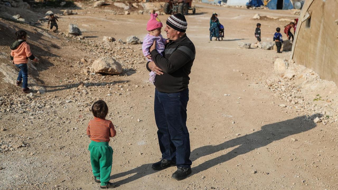 Ein Mann mit Kind auf dem Arm steht in einem staubigen Flüchtlings-Lager in Syrien. Ein Mann mit Kind auf dem Arm steht in einem staubigen Flüchtlings-Lager in Syrien.