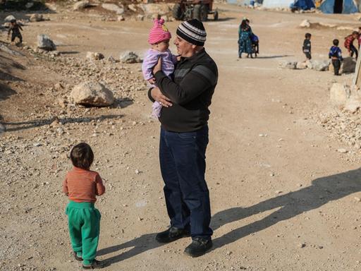 Ein Mann mit Kind auf dem Arm steht in einem staubigen Flüchtlings-Lager in Syrien.