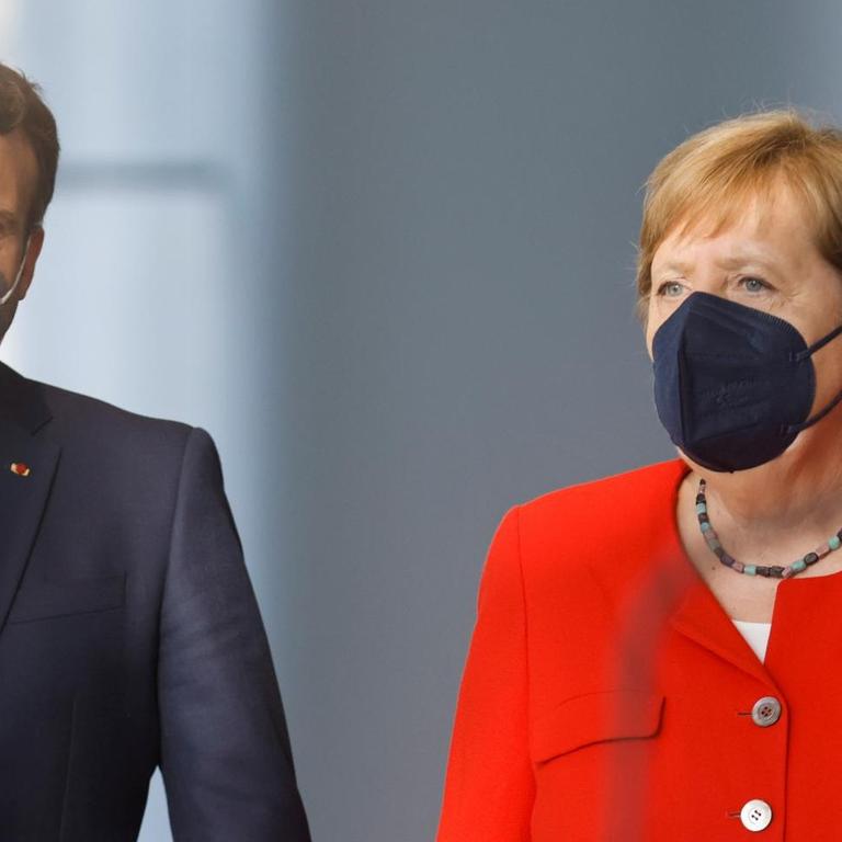 Macron trägt einen dunklen Anzug und Mund-Nase-Schutz, die Kanzlerin einen roten Blazer und einen Mund-Nase-Schutz in schwarz. Beide gehen nebeneinander her.