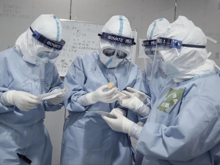 Vier Krankenschwestern im Schutzanzug betrachten Reagenzgläser mit Virenproben durch ihre Schutzbrillen.