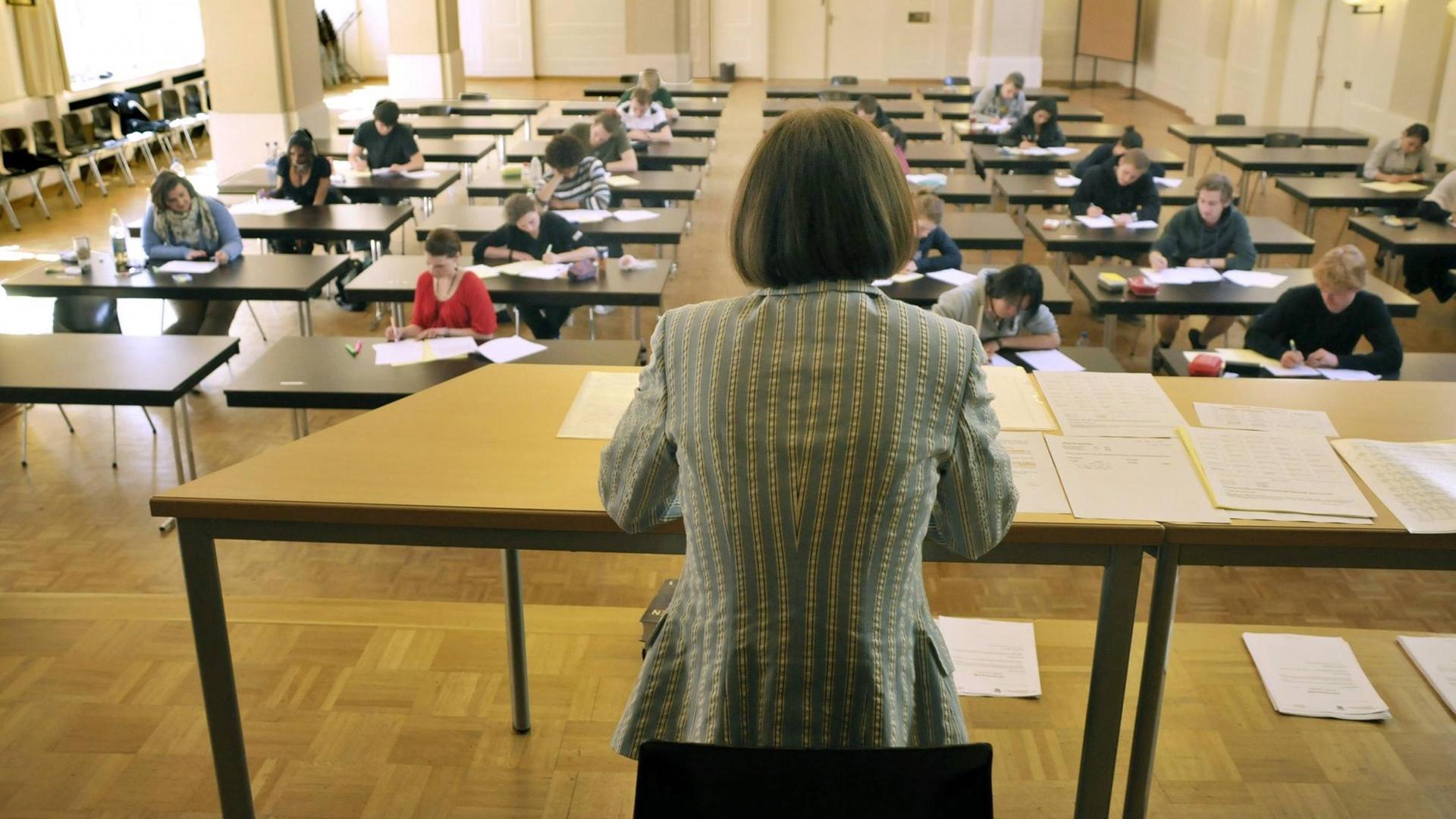 Eine Frau mit dem Rücken zum Betrachter beobachtet Schüler, die an Einzeltischen sitzen und ihr Abitur schreiben.