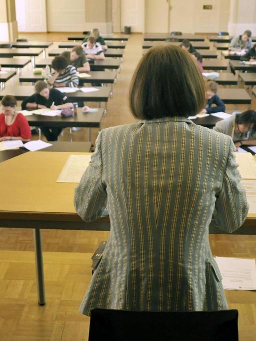Eine Frau mit dem Rücken zum Betrachter beobachtet Schüler, die an Einzeltischen sitzen und ihr Abitur schreiben.