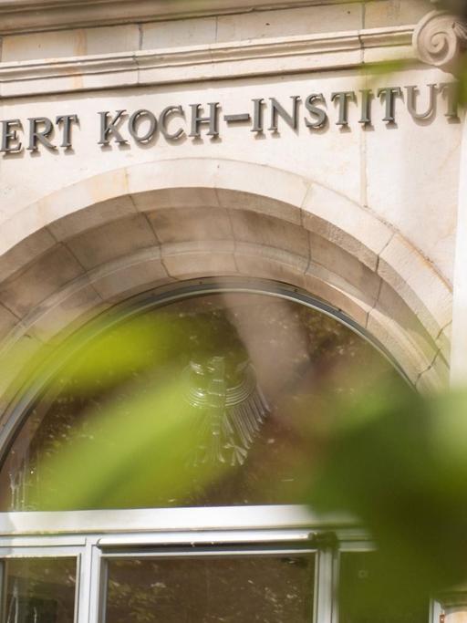 Der Eingang des Robert Koch-Instituts ist mit seinem Namenslogo durch Bäume hindurch zu sehen.