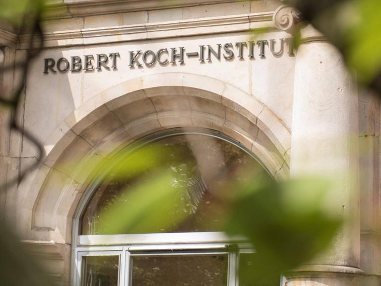 Der Eingang des Robert Koch-Instituts ist mit seinem Namenslogo durch Bäume hindurch zu sehen.