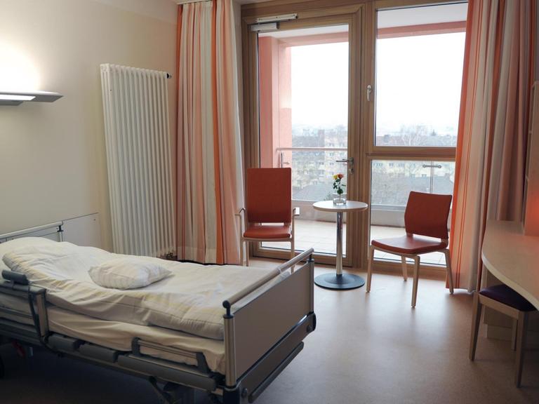 Blick in ein Patientenzimmer mit Bett, Tisch, zwei Stühlen und dem Ausblick auf einen Balkon.