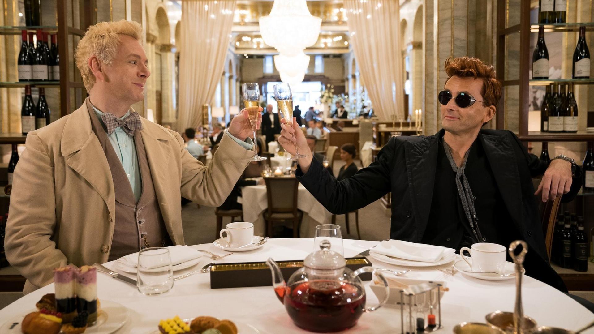 Michael Sheen (li.) als Erzengel Raphael und David Tennant als Dämon Crowley in einer Szene der Comedy-Serie "Good Omens". Die beiden sitzen an einem gedeckten Dinner-Tisch und prosten sich zu. Michael Sheen (li.) als Erzengel Raphael und David Tennant als Dämon Crowley in einer Szene der Comedy-Serie "Good Omens". Die beiden sitzen an einem gedeckten Dinner-Tisch und prosten sich zu.
