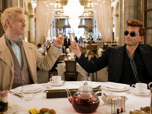 Michael Sheen (li.) als Erzengel Raphael und David Tennant als Dämon Crowley in einer Szene der Comedy-Serie "Good Omens". Die beiden sitzen an einem gedeckten Dinner-Tisch und prosten sich zu.