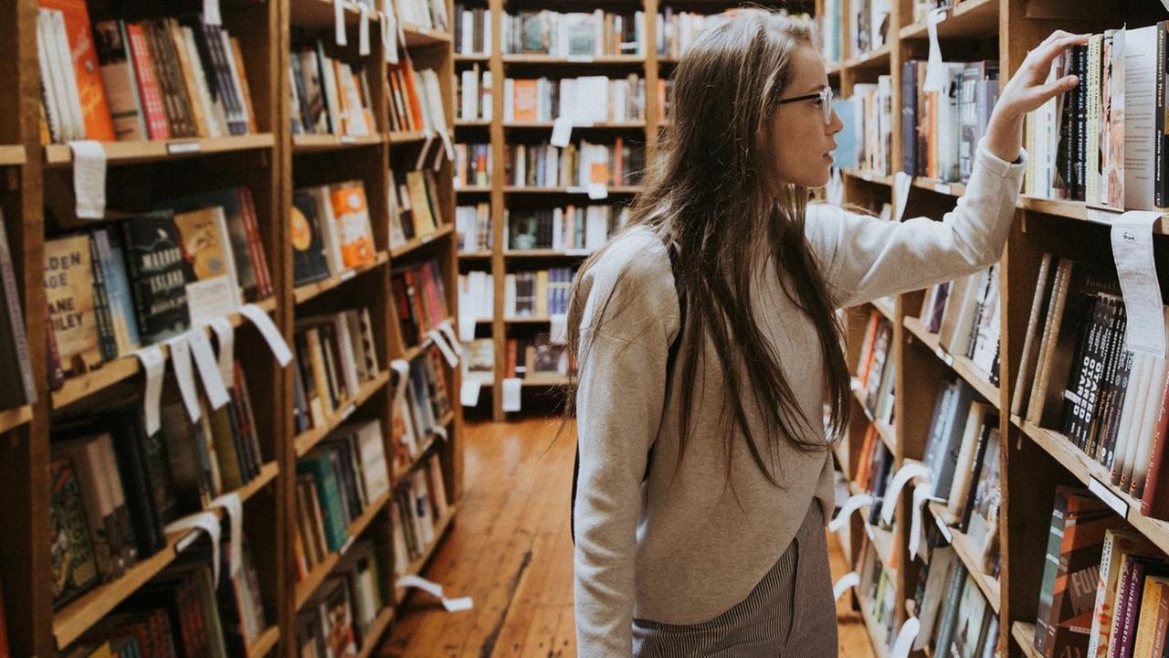 Eine junge Frau steht zwischen Bücherregalen in einem Buchladen und stöbert. Eine junge Frau steht zwischen Bücherregalen in einem Buchladen und stöbert.