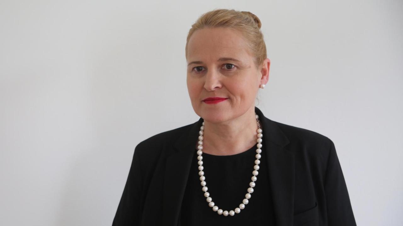 Eine Frau mittleren Alters, im schwarzen Blazer, mit Perlenkette und blondem Haarknoten, schaut versonnen an der Kamera vorbei. Eine Frau mittleren Alters, im schwarzen Blazer, mit Perlenkette und blondem Haarknoten, schaut versonnen an der Kamera vorbei.