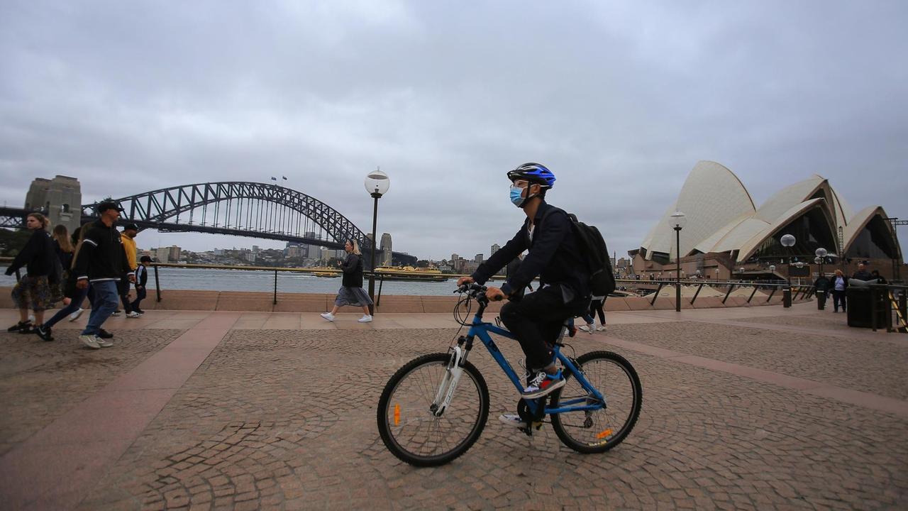 Ein Mann mit einer Atemschutzmaske fährt mit seinem Fahrrad an der Harbour Bridge und dem Opera House in Sydney vorbei.