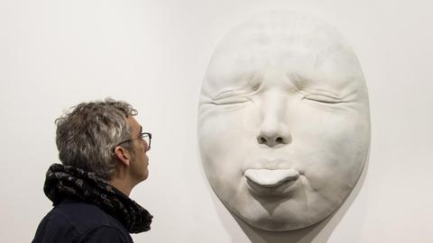 Der spanische Künstler Samuel Salcedo vor der, von ihm geschaffenen Plastik eines hyperrealistischen Gesichts, mit herausgestreckter Zunge.