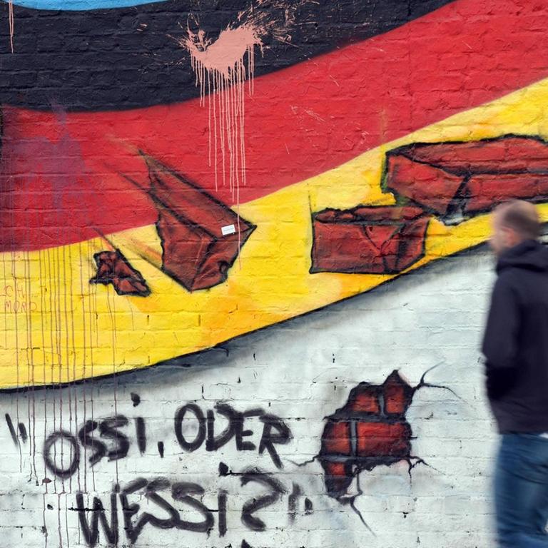 Passanten gehen an einem Wandbild von Caspar Kirchner mit der deutschen Nationalflagge und dem Schriftzug "Ossi oder Wessi? vorbei.