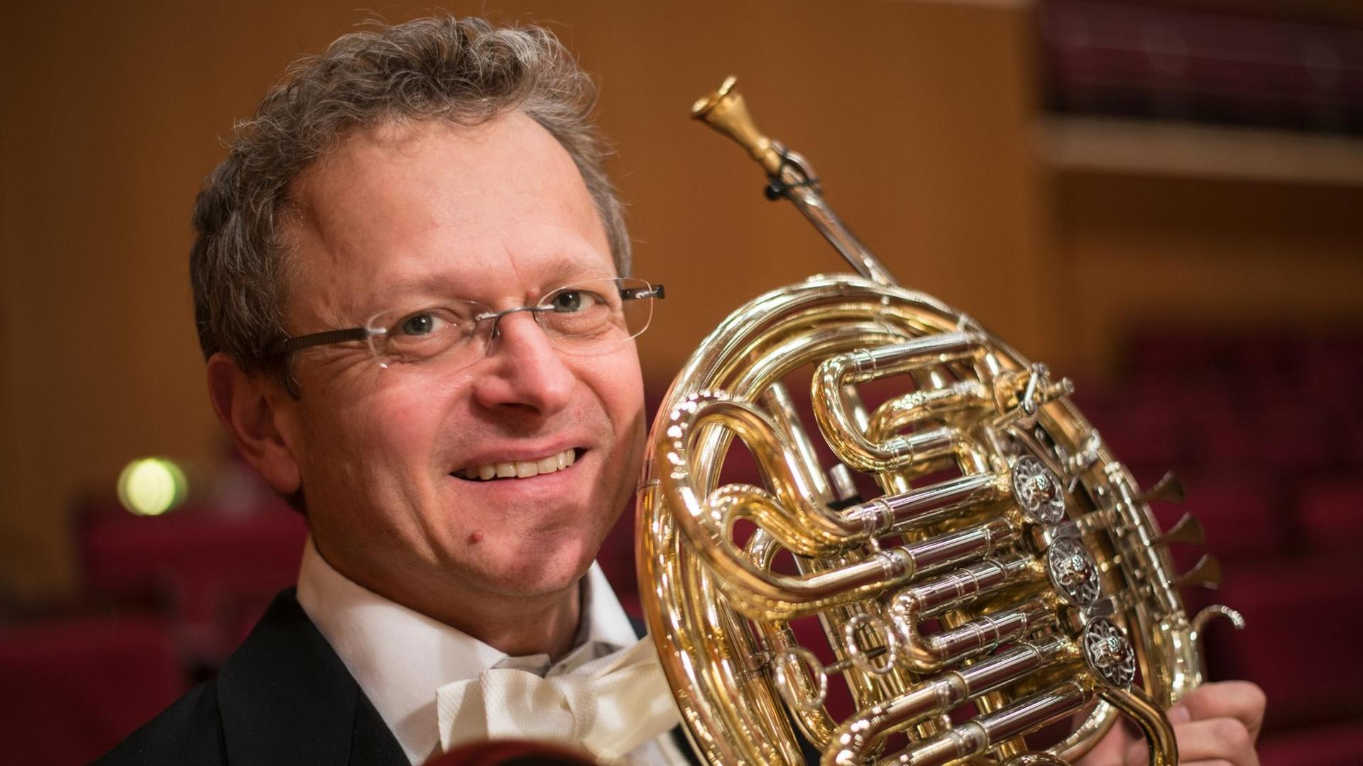 Ein Hornist mit grauen Locken und Brille, im Konzertanzug, hält sein Instrument in die Kamera. Ein Hornist mit grauen Locken und Brille, im Konzertanzug, hält sein Instrument in die Kamera.