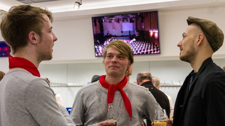 Zwei junge Männer in grauen Sweatshirts und rotem Tuch um den Hals sprechen mit dem einem dritten Mann, der Jackett und Schiebermütze trägt. Zwei junge Männer in grauen Sweatshirts und rotem Tuch um den Hals sprechen mit dem einem dritten Mann, der Jackett und Schiebermütze trägt.