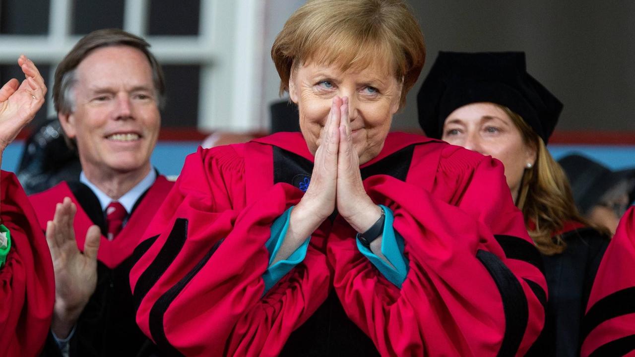 Kanzlerin Angela Merkel trägt eine rote Robe der Harvard-Universität und hat die Hände vor ihrem lächelnden Gesicht zu einer Dankesgeste zusammengelegt, nachdem sie die Ehrendoktorwürde verliehen bekam