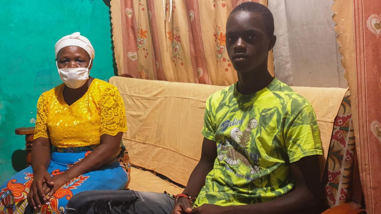 Ein afrikanischer Junge in grün gemustertem T-Shirt und Jeans sitzt neben einer älteren Frau mit Mund-Nasenschutz auf einem Sofa. Ein afrikanischer Junge in grün gemustertem T-Shirt und Jeans sitzt neben einer älteren Frau mit Mund-Nasenschutz auf einem Sofa.